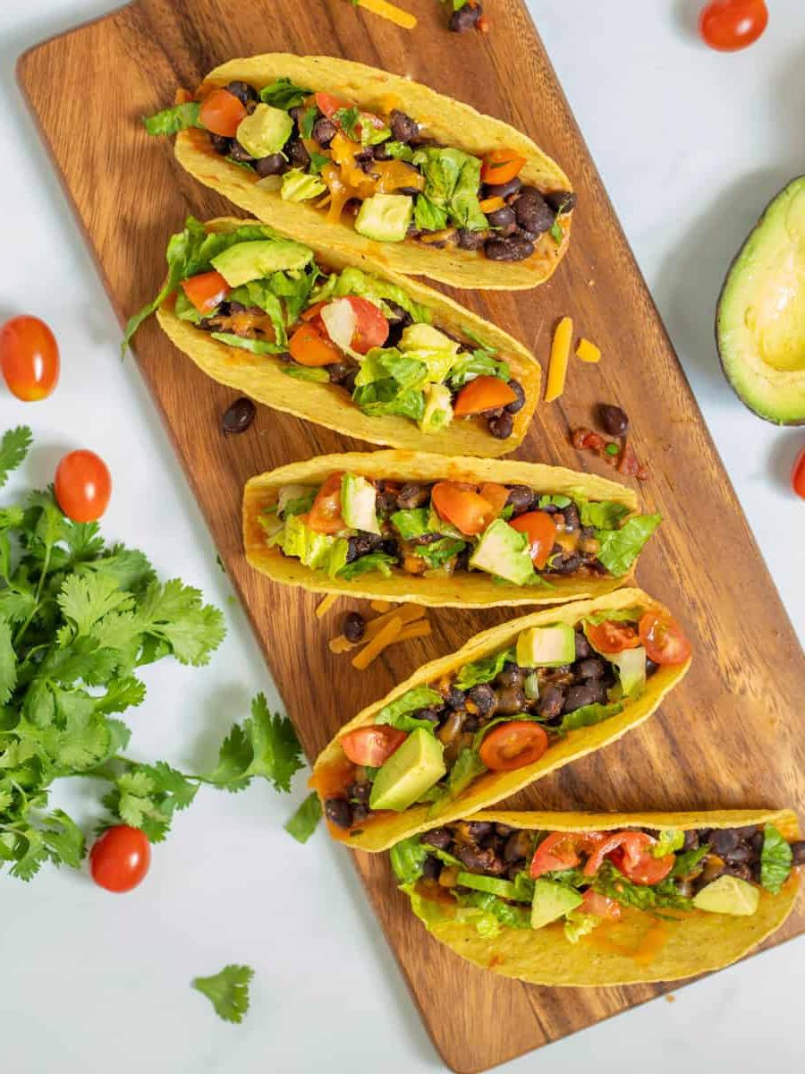 Schwarze Bohnen Tacos Schwarze-Bohnen-Tacos auf einem Holzbrett, belegt mit frischen Tomaten, Salat, Koriander und Avocado.