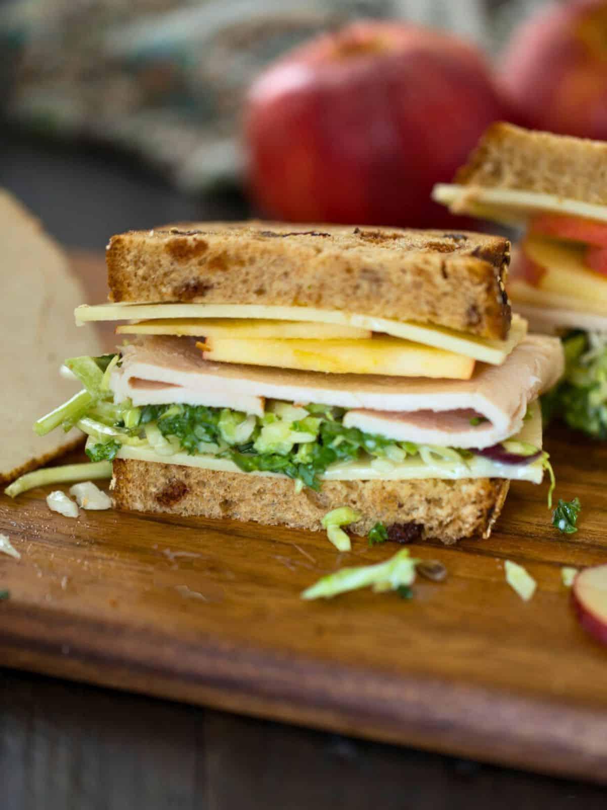 Aufgeschnittenes Truthahn-Apfel-Sandwich auf einem Schneidebrett, das Truthahnscheiben, weißen Cheddar, Grünkohl und Apfelscheiben zeigt.