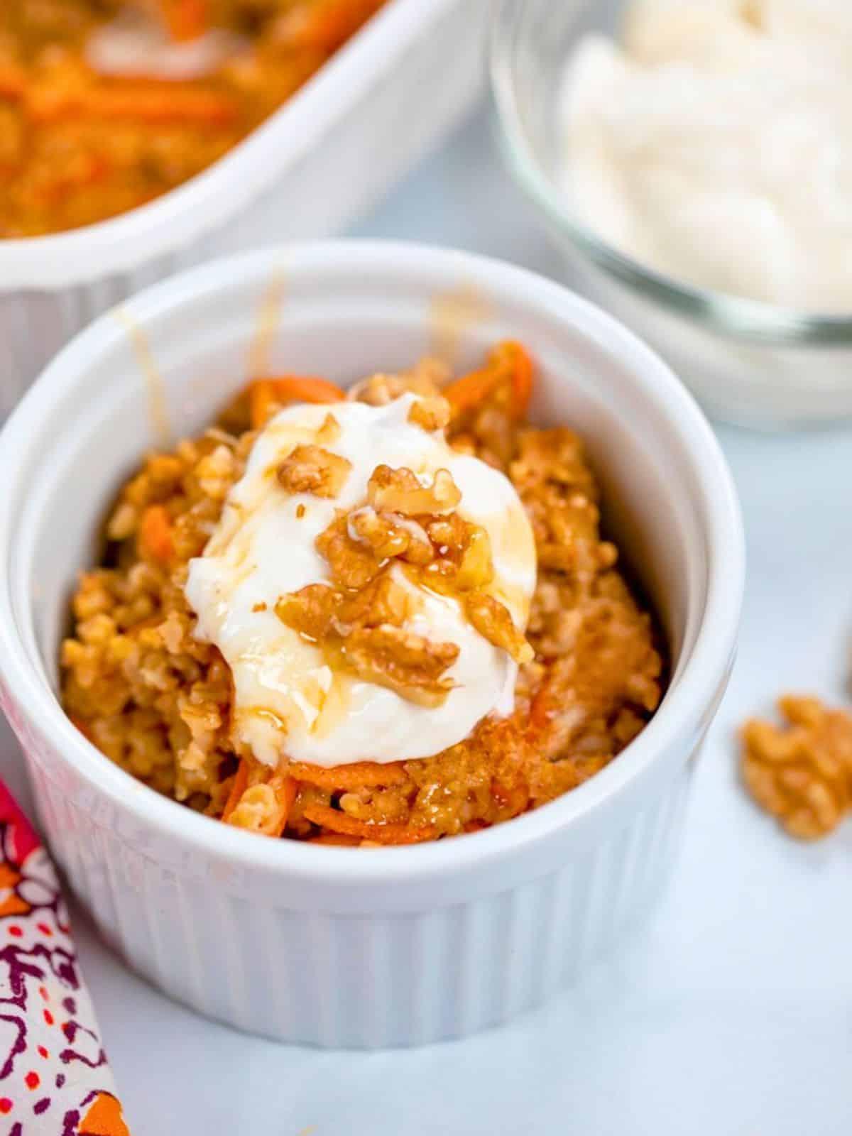 Karottenkuchen-Haferflocken-Auflauf in einer kleinen weißen Schüssel angerichtet, garniert mit griechischem Joghurt, Ahornsirup und gehackten Walnüssen.