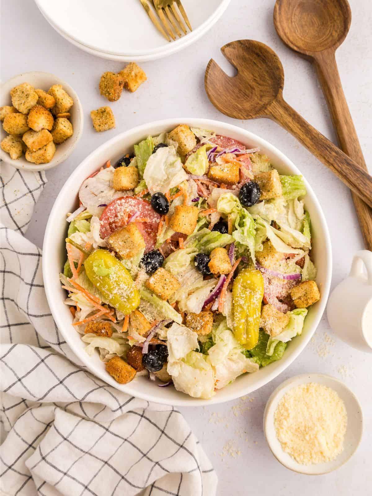 Nachgemachter Olive Garden Salat mit Eisbergsalat-Mischung, Oliven, Tomaten und Croutons in einer großen weißen Servierschüssel neben einer kleinen Schüssel mit Parmesankäse.