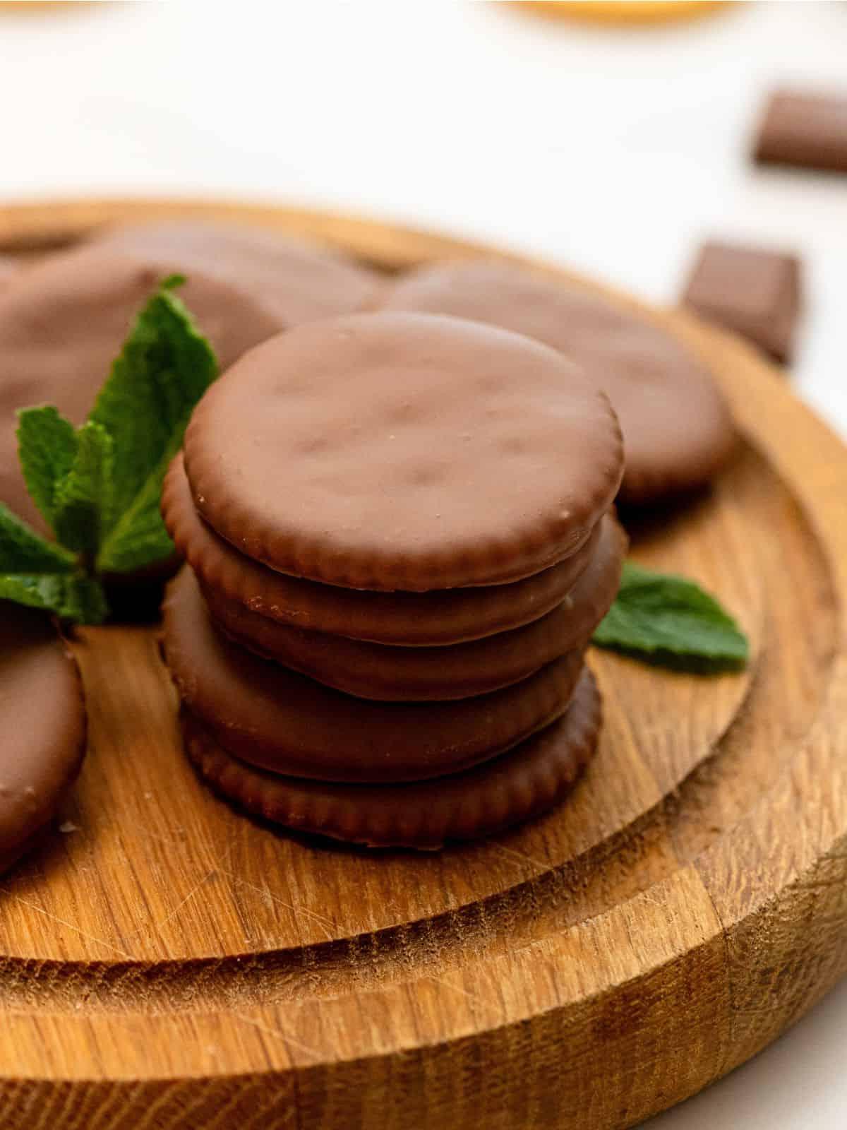 Stapel von 5 selbstgemachten Thin Mint Keksen aus runden Butterkeksen auf einem Holzschneidebrett mit frischen Minzblättern daneben.