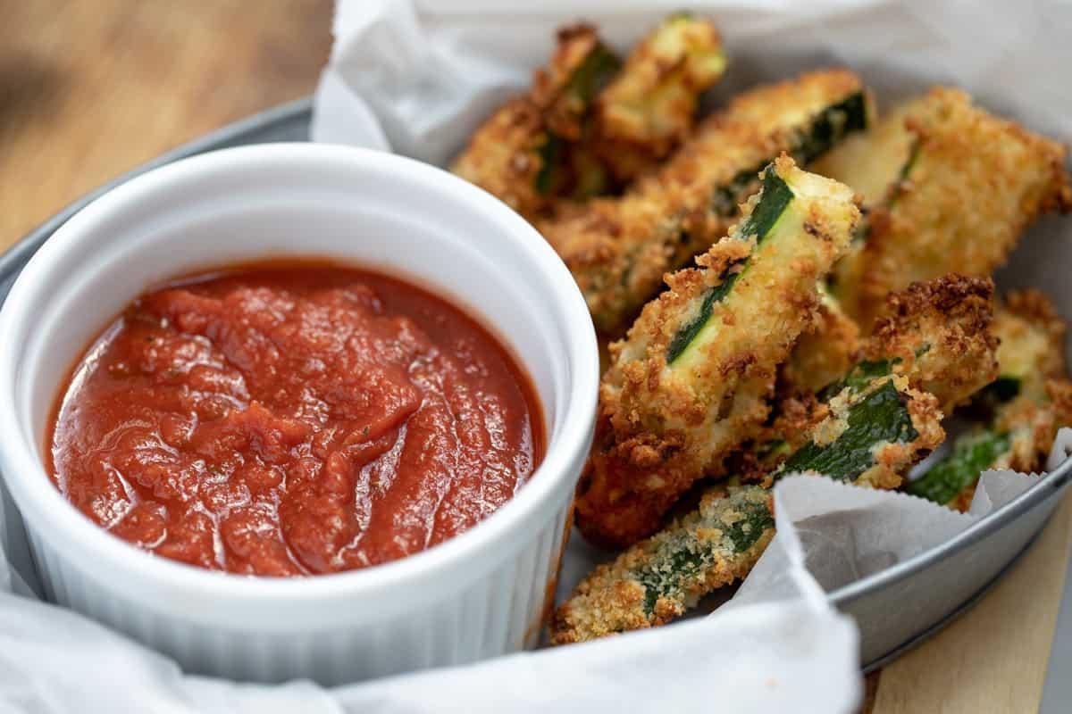 Ein Korb voller Zucchini Pommes aus der Heißluftfritteuse mit Marinara-Sauce.
