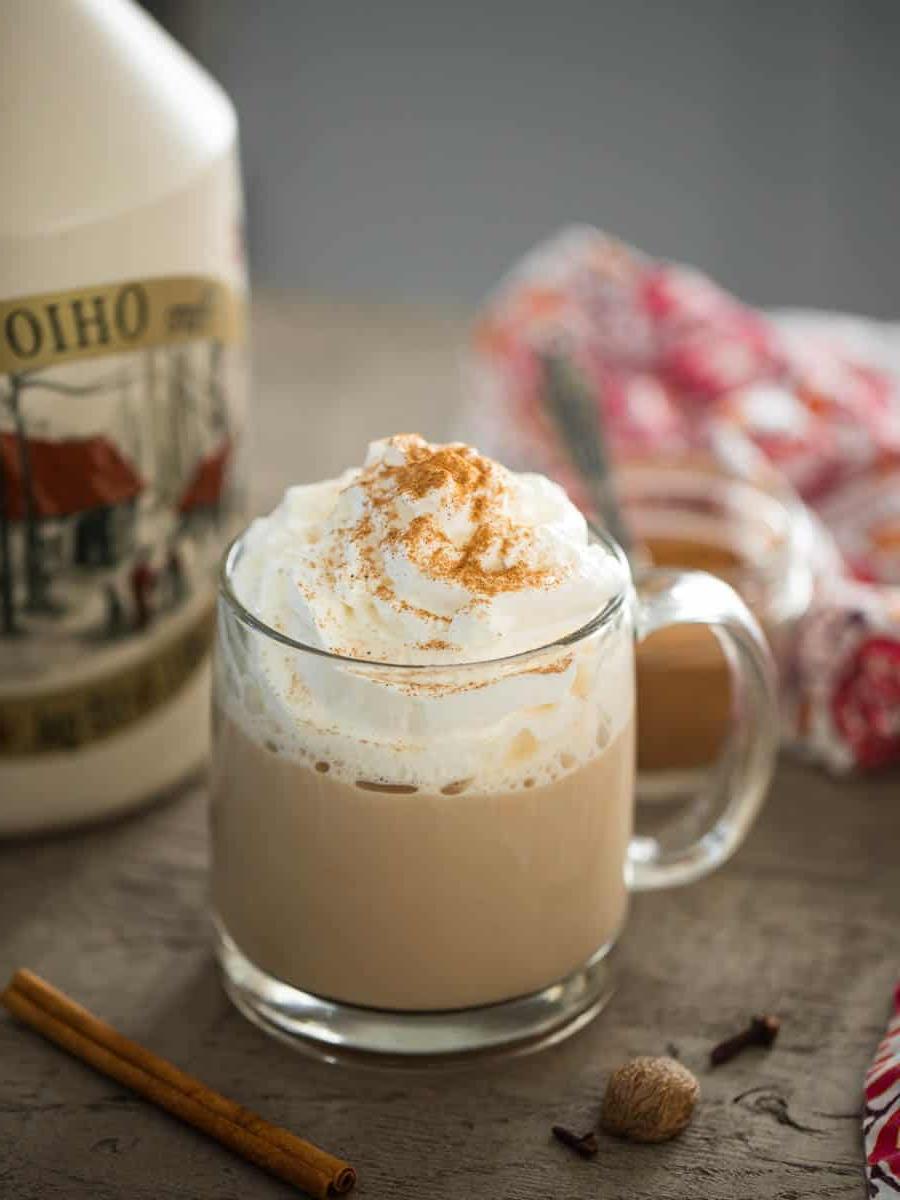 Kürbisgewürz Latte in einem durchsichtigen Glasbecher mit Ahornsirup und Zimtstangen im Hintergrund.
