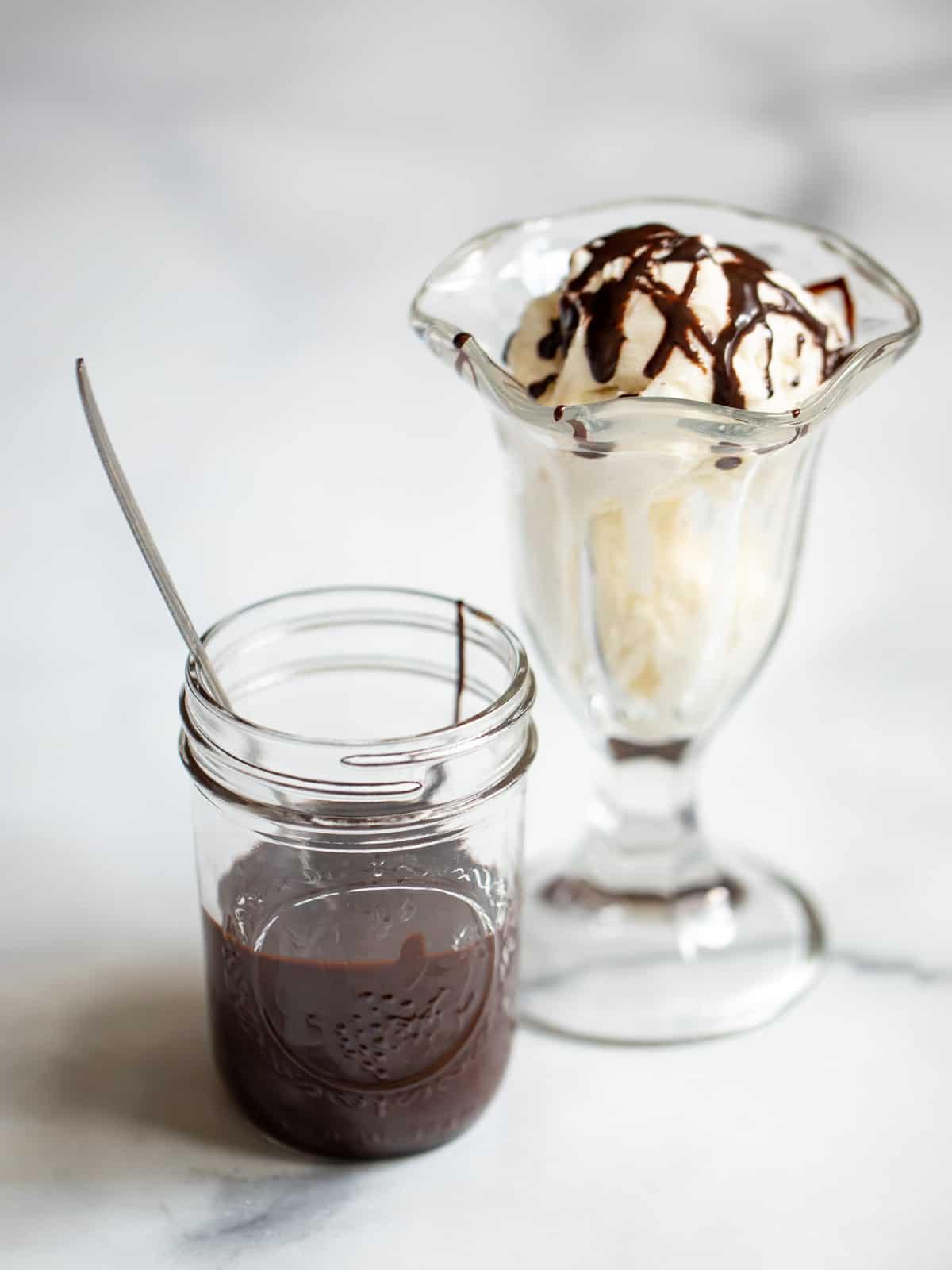 Ein Glas selbstgemachter Schokoladensirup neben einem Vanilleeisbecher mit Sahne und Kirsche.