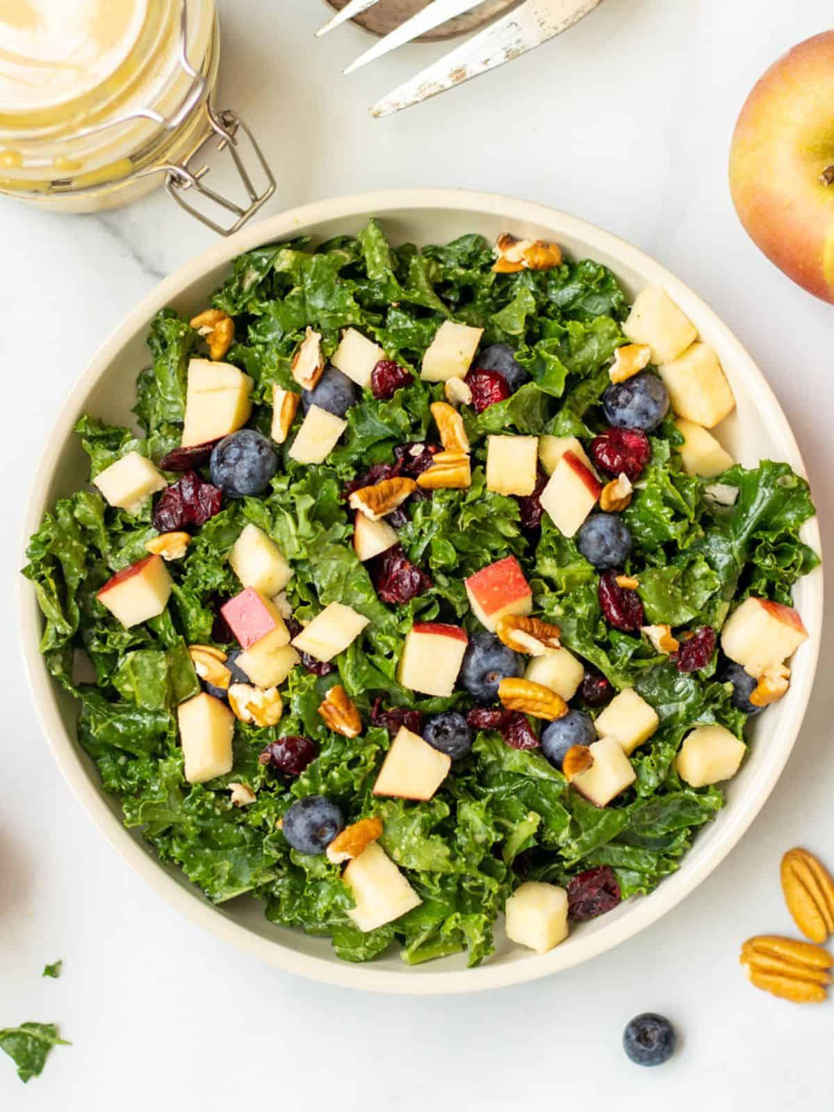 Grünkohlsalat mit Äpfeln, Beeren und Nüssen in einer Schüssel, mit Pekannüssen und Äpfeln im Hintergrund.