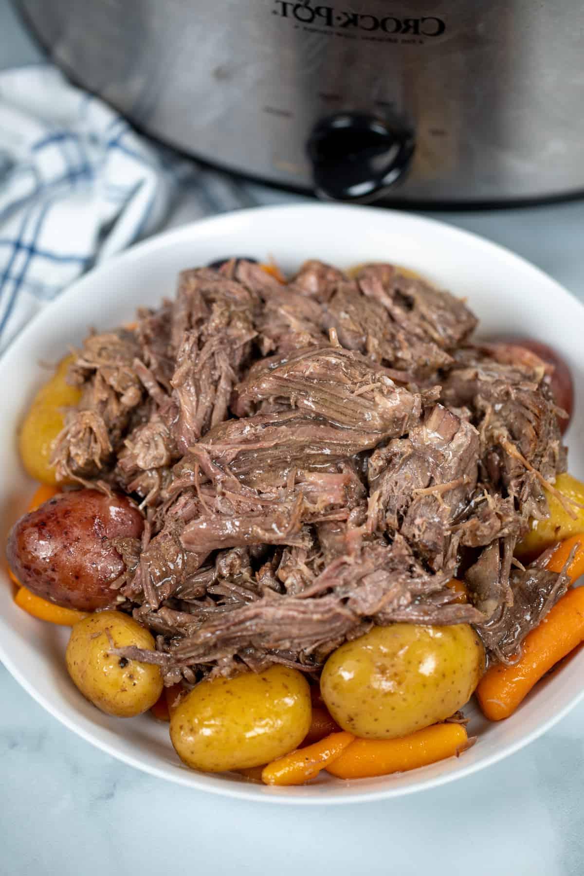 Zerkleinerter Schmorbraten in einer weißen Schale über Kartoffeln und Karotten, neben einem Slow Cooker.