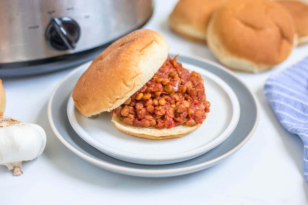 Vegane Linsen-Sloppy-Joes Vegane Sloppy Joes mit Linsen auf einem Brötchen auf einem Teller mit dem Slow Cooker im Hintergrund.