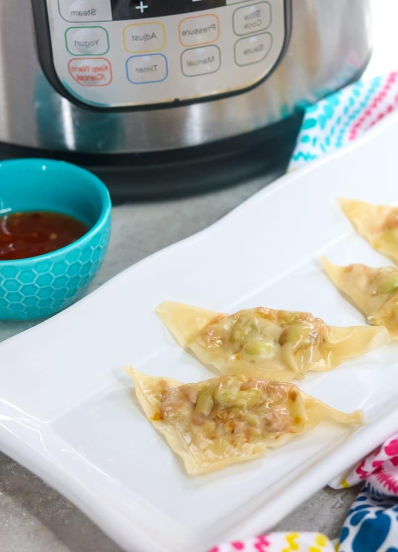 Weißes rechteckiges Tablett mit gedämpften vegetarischen Potstickers, daneben ein Instant Pot und eine blaue Schale mit hausgemachter süßer Chilisauce.