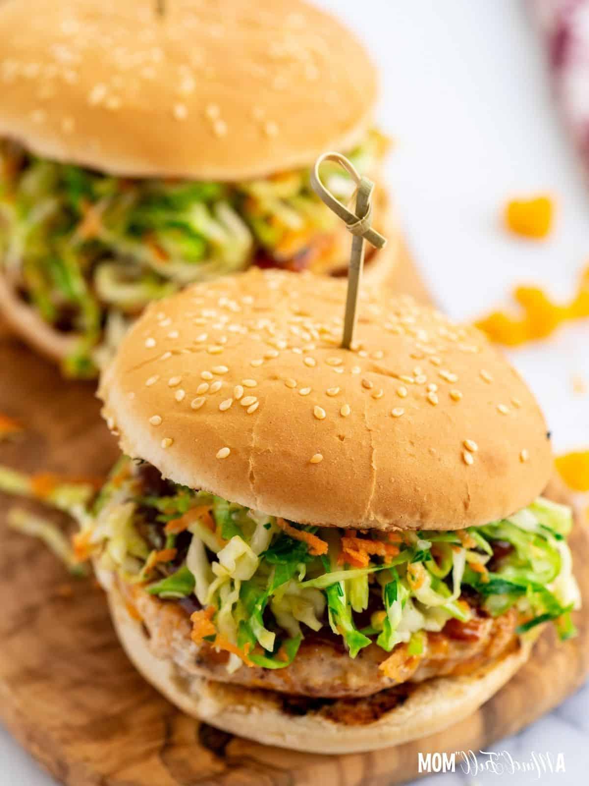 Two chicken burgers topped with coleslaw on wooden cutting board.