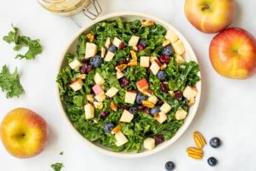 Grünkohlsalat mit Äpfeln, Beeren und Nüssen in einer Schüssel mit Pekannüssen und Äpfeln im Hintergrund.