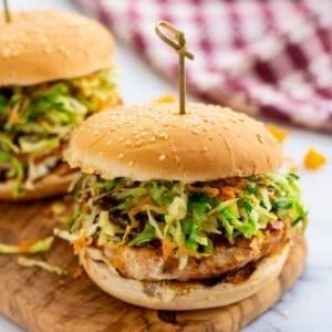 Juicy chicken burger on bun topped with BBQ sauce and coleslaw on cutting board.