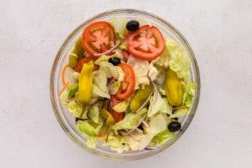 Klare Rührschüssel mit Eisbergsalat, Kohl, Zwiebeln, Karotten, Oliven, Tomaten und Pepperoncini-Paprika vermischt.