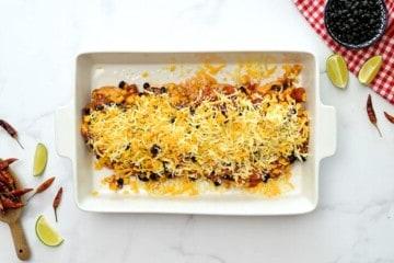 Gewürzte Hähnchenbrustfilets, bedeckt mit Bohnen, Mais, Tomaten und geriebenem Käse in einer weißen Auflaufform.