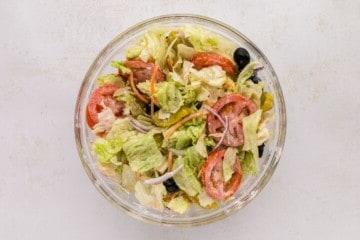 Eisbergsalat, Kohl, Zwiebeln, Karotten, Oliven, Tomaten und Pepperoncini-Paprika und hausgemachtes cremiges italienisches Dressing in einer klaren Rührschüssel nach dem Vermischen.