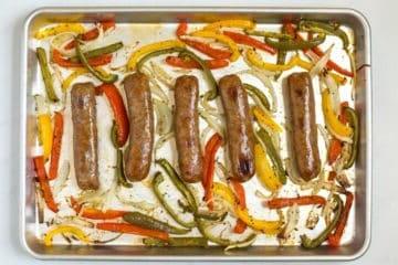 5 gebackene Würste auf einem Backblech mit Rand, umgeben von Zwiebeln und Paprika.
