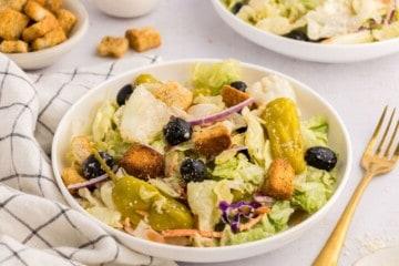 Nachgemachter Olive Garden Salat mit Eisbergsalat-Mischung, Oliven, Tomaten und Croutons in einer großen weißen Servierschüssel neben einer Gabel.