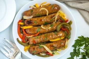 Gebackene italienische Wurst in einer weißen Servierschale, garniert mit Paprika, Zwiebeln und gehackter Petersilie.