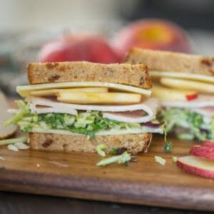 Aufgeschnittenes Truthahn-Apfel-Sandwich auf einem Schneidebrett, das Truthahnscheiben, weißen Cheddar, Grünkohl und Apfelscheiben zeigt.