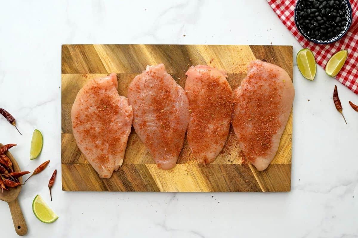 Cowboy Hähnchen Auflauf Rezept Vier entbeinte Hähnchenbrustfilets ohne Haut auf einem Holzschneidebrett, gewürzt mit Tex-Mex-Gewürzen.