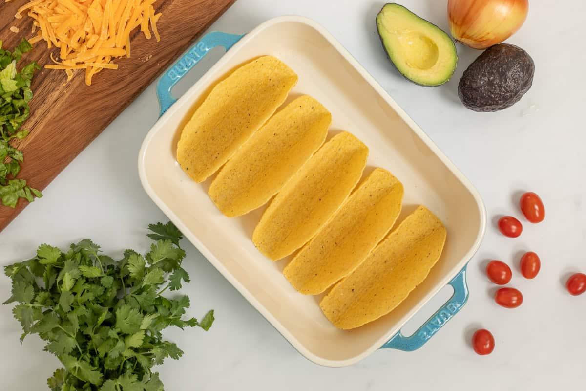 Einfaches Schwarze-Bohnen-Taco-Rezept Taco-Schalen in einer Auflaufform neben frischem Koriander, Avocado und Käse.