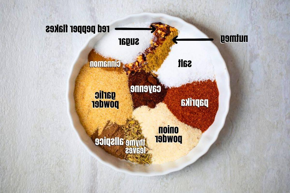 Zutaten für Jerk Gewürz Zucker, Salz, Zwiebelpulver, Knoblauchpulver, Thymianblätter, Piment, rote Pfefferflocken, Zimt, Muskatnuss, Paprika, Cayennepfeffer und Zucker, beschriftet in Schalen zur Herstellung von hausgemachtem Jerk Gewürz.