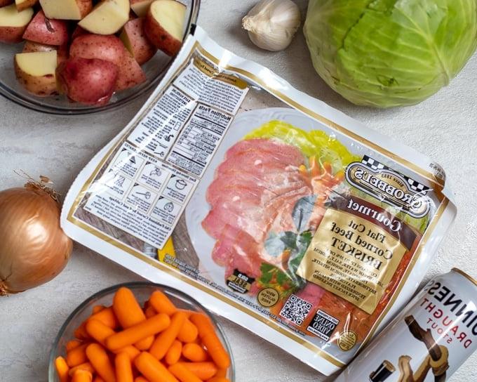 Zutaten für Corned Beef und Kohl auf einem weißen Brett: Corned Beef, Kohl, Zwiebel, rote Kartoffeln, Karotten und Bier.