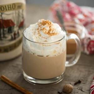 Kürbisgewürz Latte in einem durchsichtigen Glasbecher mit Ahornsirup und Zimtstangen im Hintergrund.