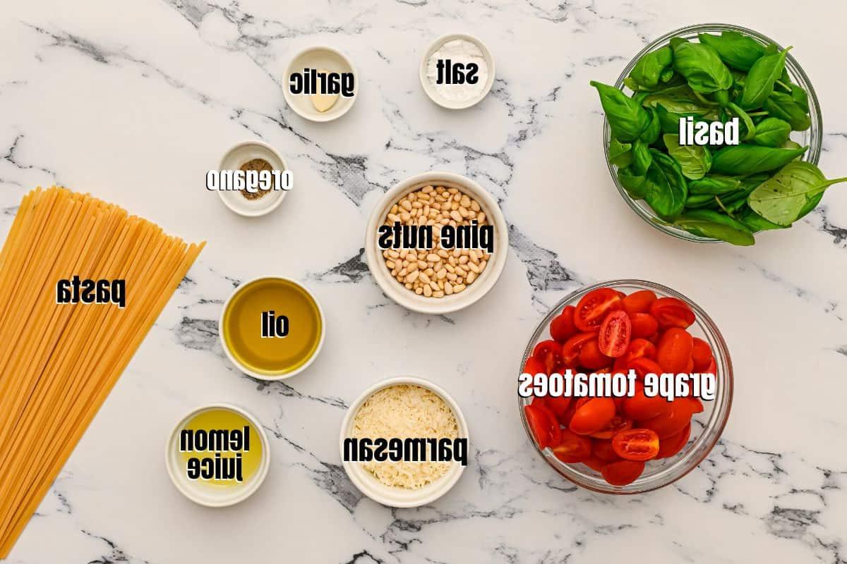 Zutaten für Pesto Nudeln auf einer Küchentheke arrangiert und beschriftet: frische Tomaten, Basilikum, Parmesan, Pasta und Olivenöl.
