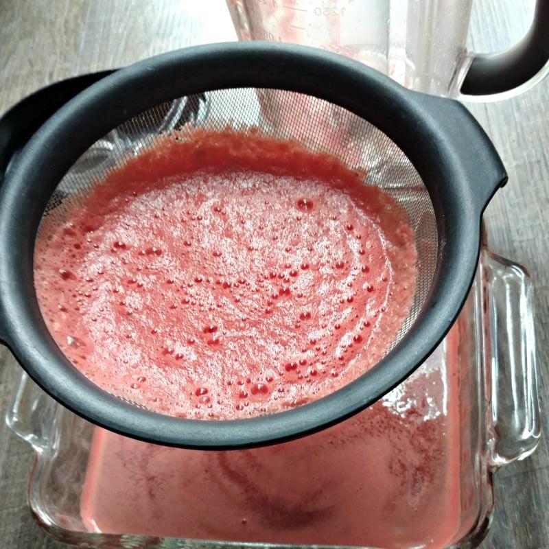 Wie man Wassermelonen Granitas macht Feinmaschiges Sieb mit Wassermelonensaft über einer Glasbackform.