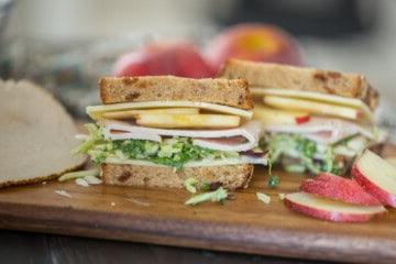 Aufgeschnittenes Truthahn-Apfel-Sandwich auf einem Schneidebrett, das Truthahnscheiben, weißen Cheddar, Grünkohl und Apfelscheiben zeigt.