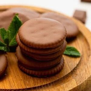 Stapel von 5 selbstgemachten Thin Mint Keksen aus runden Butterkeksen auf einem Holzschneidebrett mit frischen Minzblättern daneben.