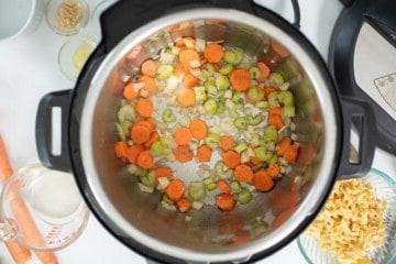 Karotten, Zwiebeln, Sellerie und Knoblauch werden im Schnellkochtopf mit Olivenöl und Butter angebraten. Zutaten für die Puten-Nudelsuppe sind im Hintergrund zu sehen.