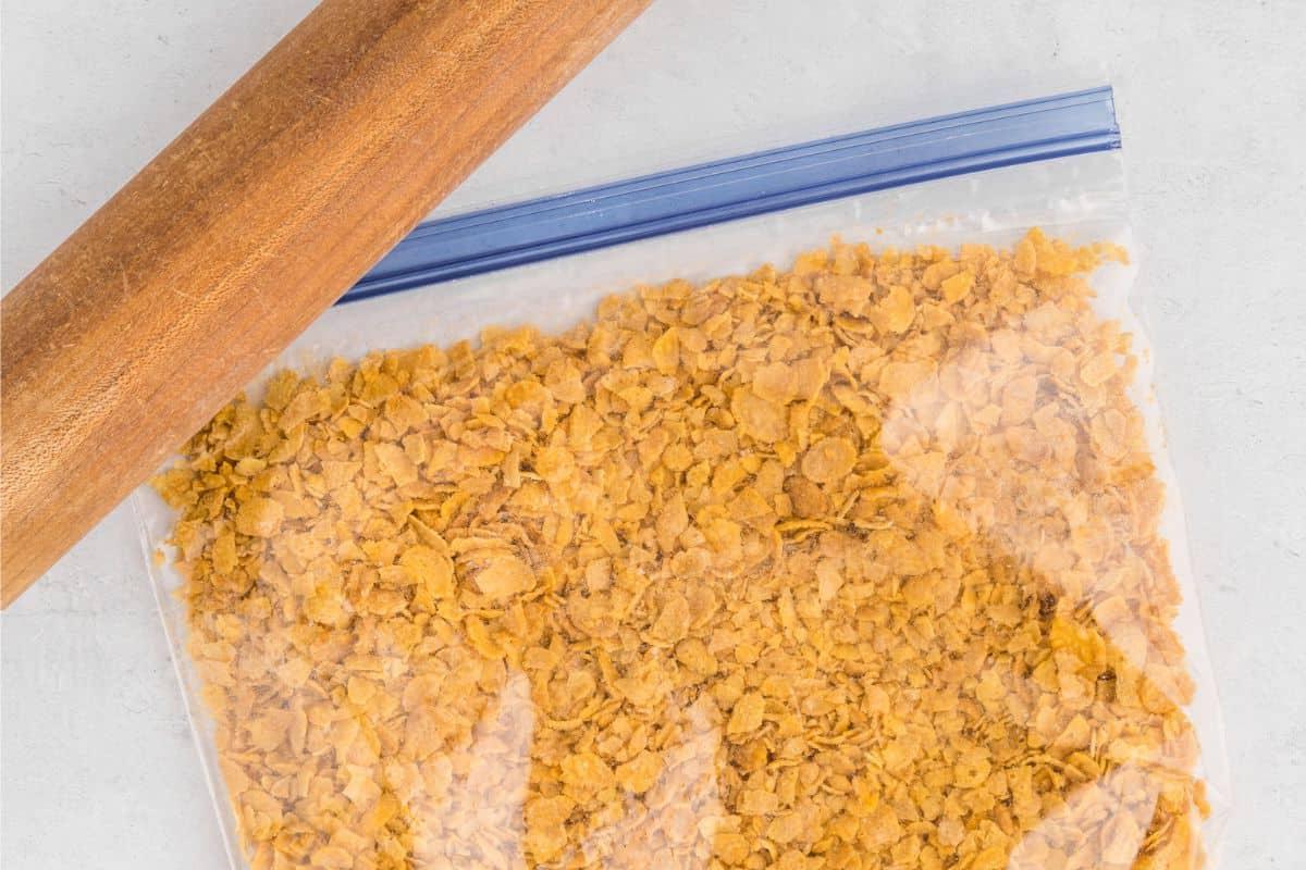 Cornflake Funeral Potatoes Hash Browns in einem Beutel mit Nudelholz daneben, nachdem die Cornflakes leicht zerkleinert wurden.
