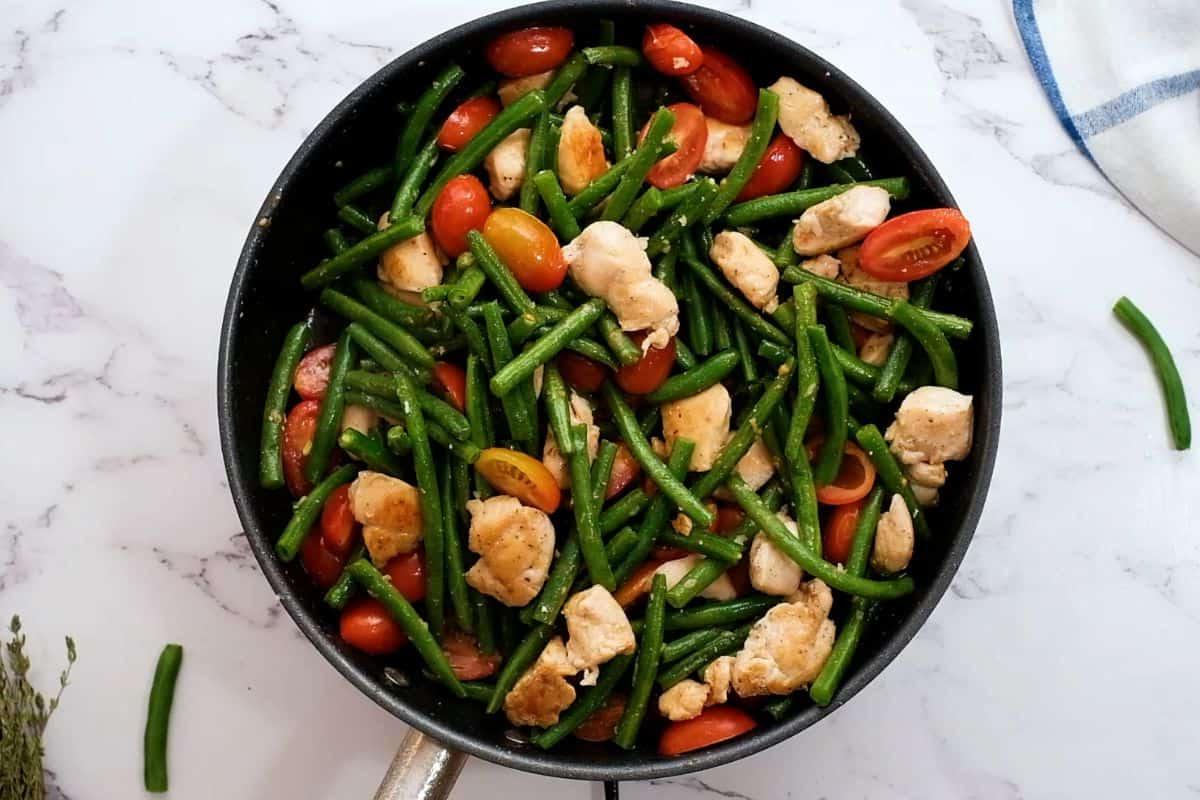 Grüne Bohnen und Tomaten zu gebratenem Hähnchen in der Pfanne mit frischem Thymian im Hintergrund hinzugefügt.