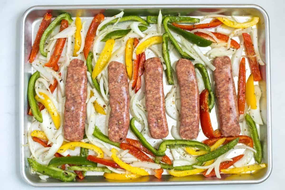 5 rohe italienische Würste auf einem Backblech mit Rand, umgeben von Zwiebeln und Paprika, gewürzt mit italienischer Kräutermischung und Knoblauch.
