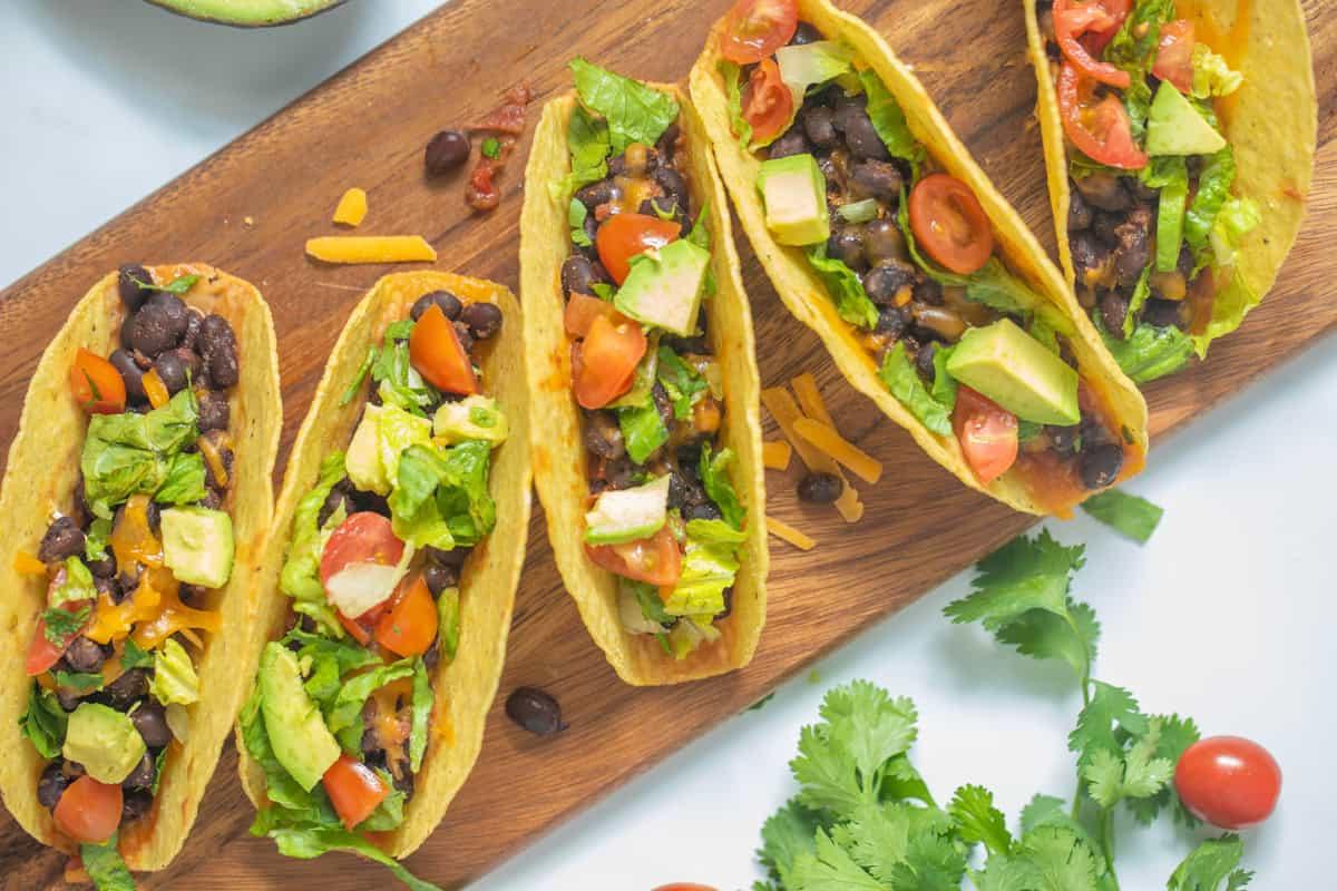 Schwarze Bohnen Taco Rezept Schwarze-Bohnen-Tacos auf einem Holzbrett, belegt mit frischen Tomaten, Salat, Koriander und Avocado.