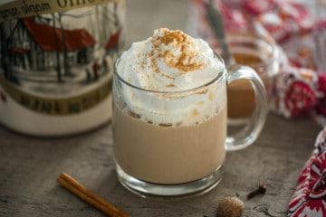 Kürbisgewürz Latte in einem durchsichtigen Glasbecher mit Ahornsirup und Zimtstangen im Hintergrund.
