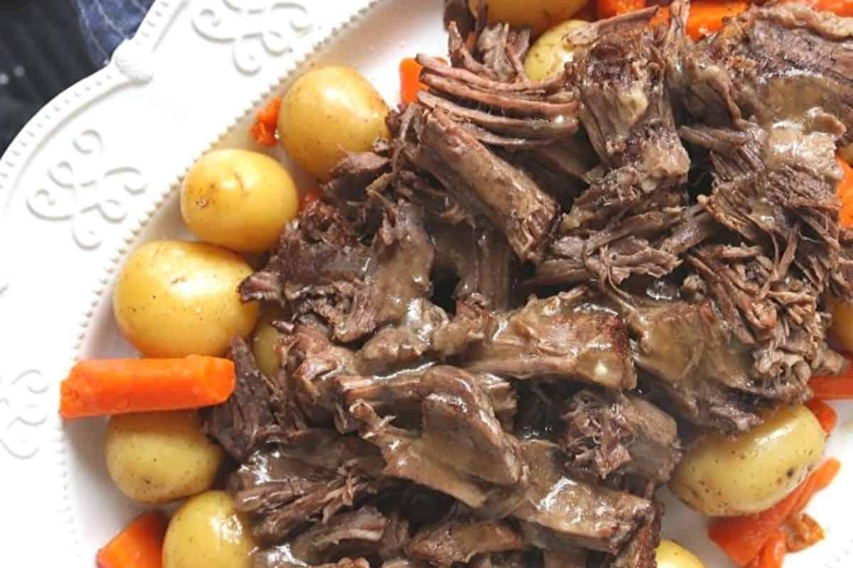 Ein dampfender Schmorbraten auf einer weißen Servierplatte, begleitet von Kartoffeln und Karotten, direkt neben dem Instant Pot.