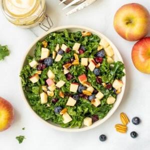 Grünkohlsalat mit Äpfeln, Beeren und Nüssen in einer Schüssel mit Pekannüssen und Äpfeln im Hintergrund.