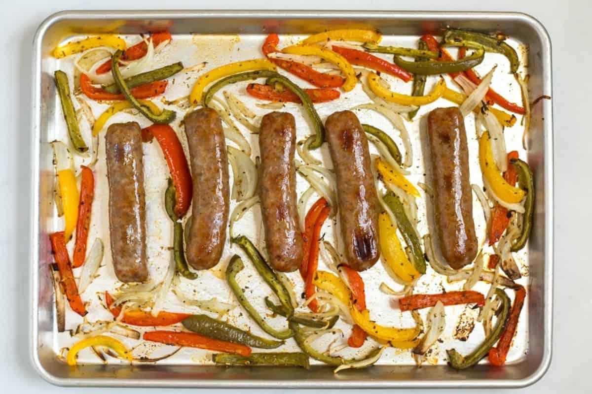 5 gebackene Würste auf einem Backblech mit Rand, umgeben von Zwiebeln und Paprika.