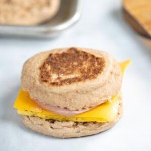 Ein nachgemachtes Egg McMuffin Frühstückssandwich auf der Theke mit getoasteten English Muffins im Hintergrund.
