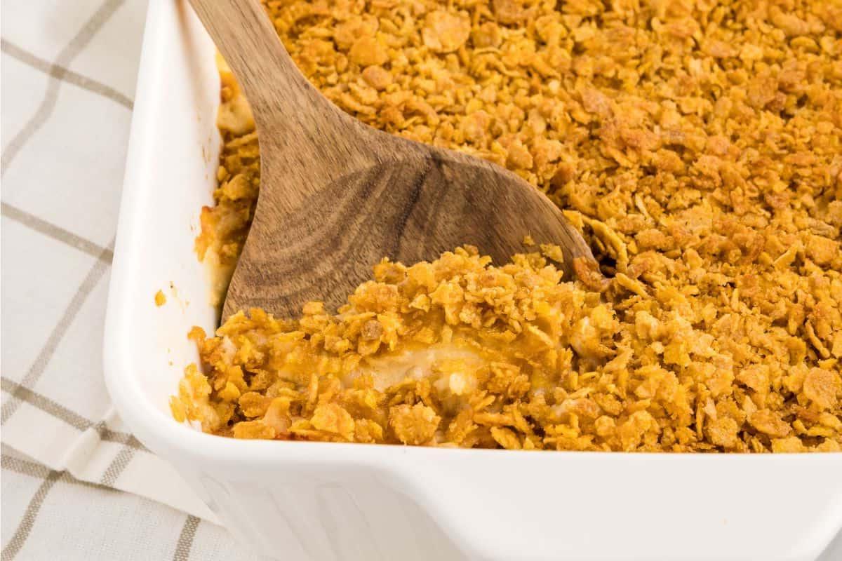 Käsige Hash-Brown-Funeral-Potatoes Holzlöffel schöpft Funeral Potatoes aus einer großen weißen Auflaufform.
