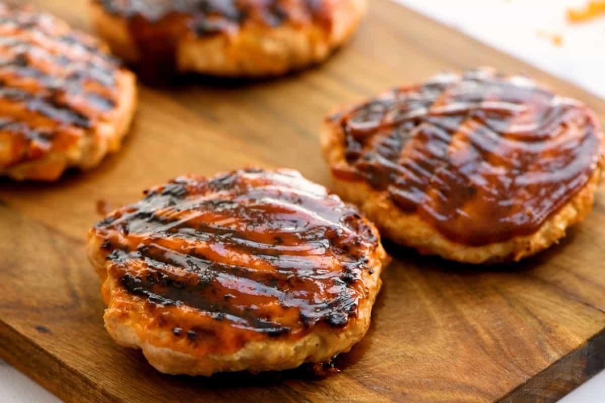 Four chicken burgers on wooden platter basted in BBQ sauce.
