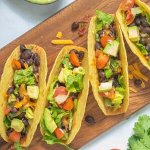Einfache Schwarze Bohnen Tacos 4 Schwarze-Bohnen-Tacos auf einem Holzbrett, belegt mit frischen Tomaten, Salat, Koriander und Avocado.