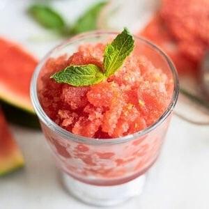 Wassermelonen-Granita 4 Wassermelonen Granita in einer Glasschale, garniert mit frischer Minze und frischer Wassermelone im Hintergrund.