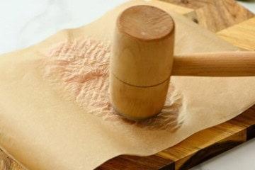 Fleischklopfer, der Hähnchenbrust zwischen 2 Stücke Backpapier klopft.