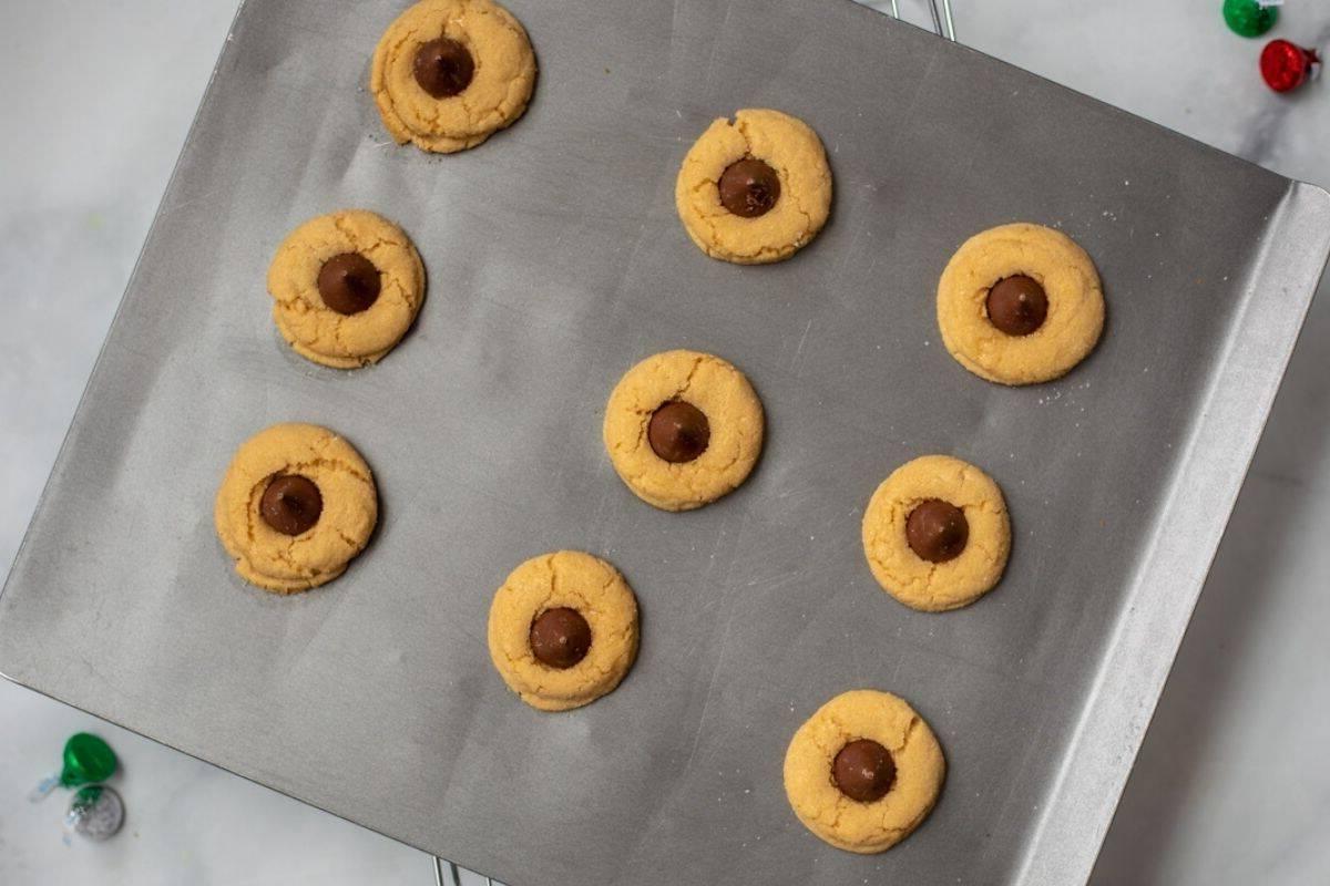 9 gebackene, weiche Erdnussbutter-Kekse, nachdem ein Schokokuss auf sie gelegt wurde, auf einem Backblech mit Hershey's Kisses im Hintergrund.