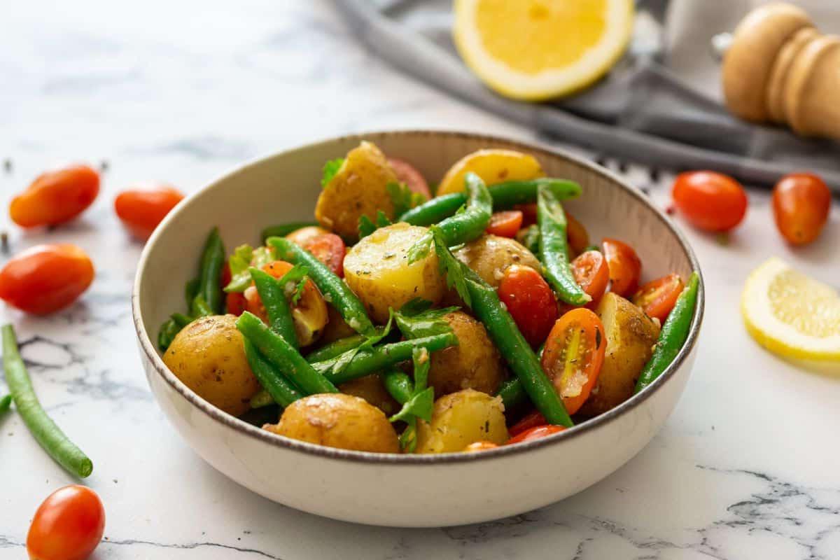 Kartoffelsalat ohne Mayo Italienischer Kartoffelsalat in einer Schüssel, garniert mit frischer Petersilie, mit Zitrone und Tomaten im Hintergrund.