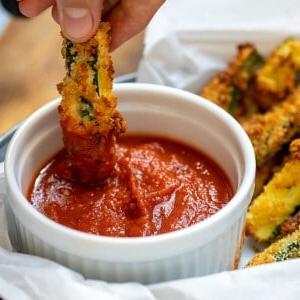 Eine Zucchini Pommes aus der Heißluftfritteuse wird in Marinara-Sauce getaucht.
