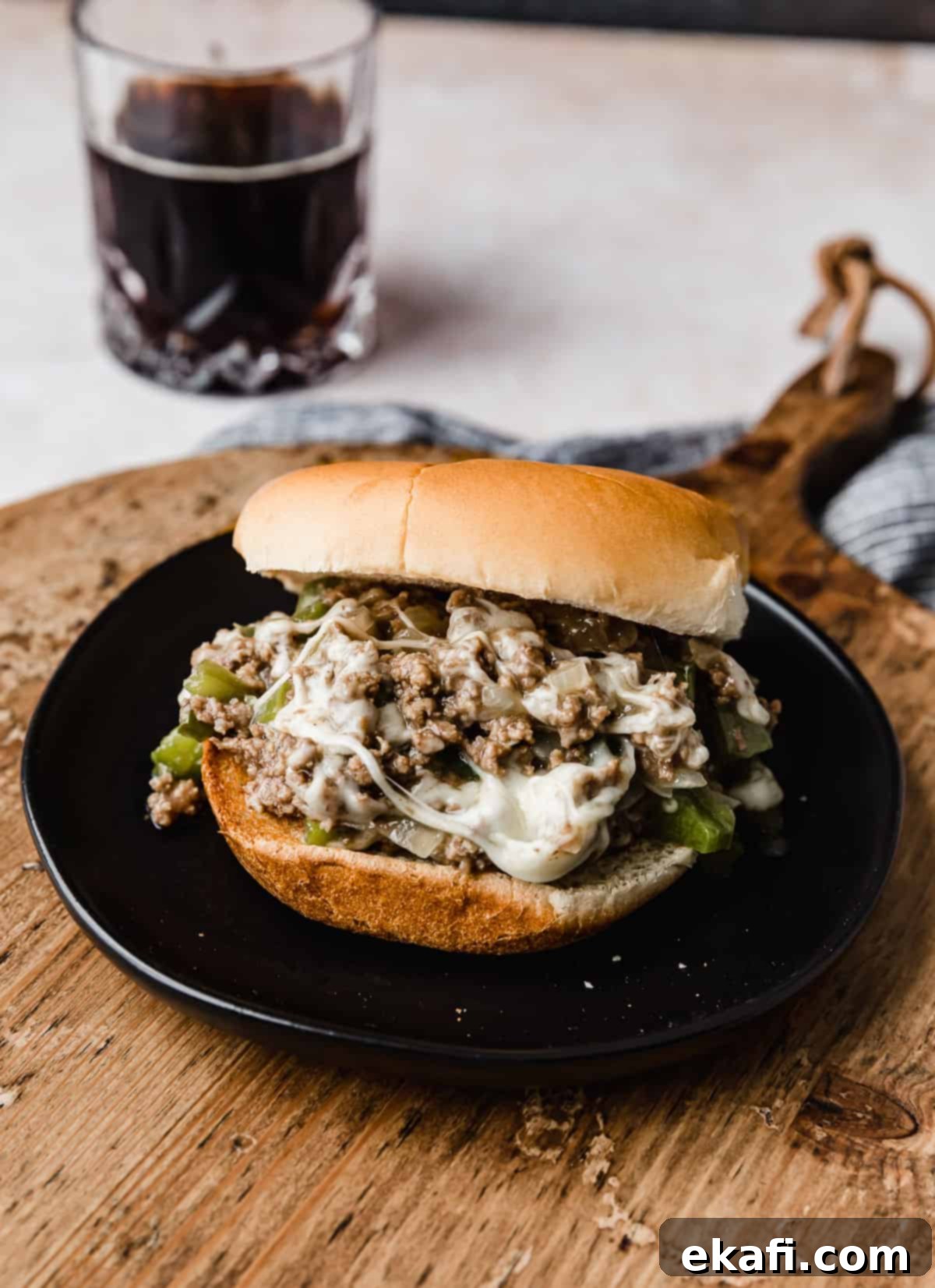 Φιλαδέλφειας Τυρόστεικ Σλόπι Τζο 2 A Philly Cheesesteak Sloppy Joe on a black plate on a wooden round cutting board.